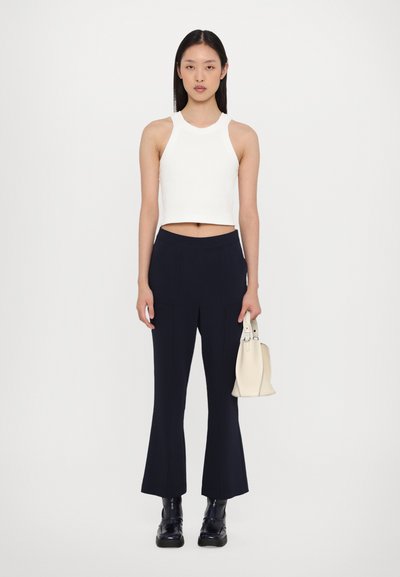 Weißes geripptes Cropped-Top, navyfarbene Schlaghose, schwarze Plateaustiefel, hält eine kleine cremefarbene Handtasche. Einfarbiger Hintergrund.