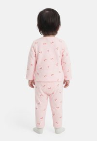 Παιδάκι που στέκεται με την πλάτη, φορώντας ροζ πυτζάμες με μικρά κόκκινα λουλουδένια σχέδια και ανοιχτόχρωμες κάλτσες σε λευκό φόντο.