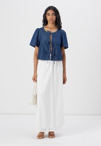 Top en denim court avec des manches bouffantes, fermeture devant ouverte, associé à une longue jupe en dentelle blanche et des sandales beiges. Petite pochette blanche en accent.