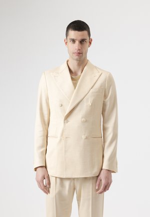 Hombre con blazer cruzado color crema y pantalones a juego, de pie contra un fondo claro y liso, mirando hacia adelante.