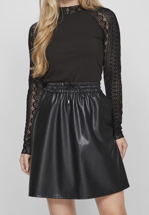 Vrouw draagt een zwarte geribde top met lange kanten mouwen en een hoog getailleerde zwarte rok van nep leer met een elastische taillekoord.