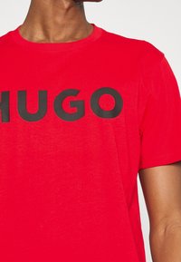 Camiseta roja de manga corta de algodón, con un destacado logo negro "HUGO" en el pecho. Cuello redondo con textura suave.