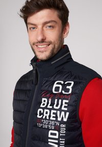 Gilet trapuntato blu navy con colletto alto, zip frontale e testo stampato in bianco e rosso. Contrasto con una camicia rossa sotto.