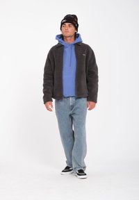 Chaqueta gris oscuro difusa sobre un hoodie azul, combinada con vaqueros azul claro y zapatillas negras. El gorro es negro con acentos coloridos.