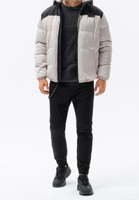 Puffer jacket in beige and black, featuring a zip closure, hood, and elastic cuffs. Paired with black pants and black sneakers.