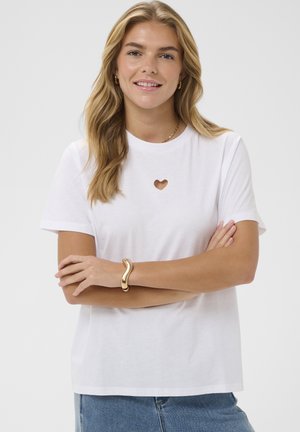 Jeune femme aux longs cheveux blonds portant un t-shirt blanc avec une découpe en forme de cœur sur la poitrine, un bracelet en or et un jean bleu, souriante.