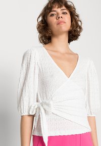 Top branco texturizado envolvente com mangas curtas em formato de puff e detalhe de cintura amarrada, combinado com calças rosas. Apresenta um decote em V profundo.