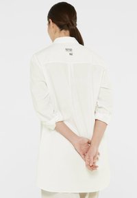 White, long-sleeve shirt with rolled cuffs, features a center seam and a label on the back neck. Smooth, lightweight fabric.
