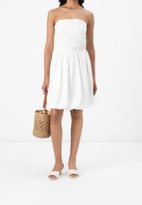 Bílé šaty bez ramínek s texturovaným živůtkem a nabíranou sukní. Pletená košíková taška s koženým uchycením a bílé nazouvací sandály se zlatými detaily.