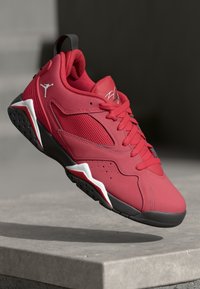Zapatilla deportiva roja y negra con acentos blancos, paneles de malla, cordones y el logo de Jumpman, posicionada sobre una superficie de concreto.