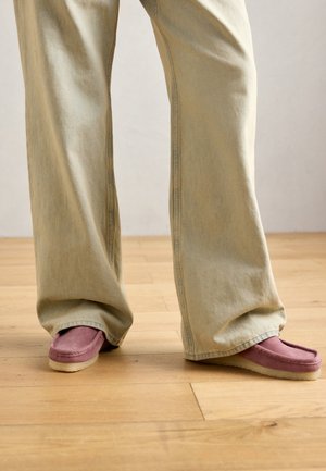 Persoon in losse beige broek en paarse suède mocassin-schoenen die op een licht houten vloer staat.