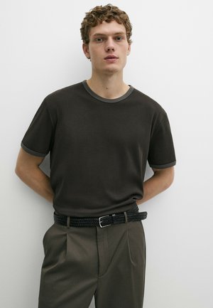 Giovane uomo con capelli ricci che indossa una camicia grigio scuro a maniche corte e pantaloni verde oliva con cintura nera intrecciata.
