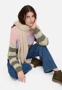 Gestreepte gebreide trui in pastelroze, lavendel en olijfgroen, met een beige sjaal, gecombineerd met flared blauwe jeans en donkergroene enkellaarzen.