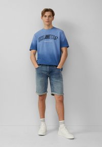 Blauwe gradient T-shirt met de tekst "ONBEPERKTE VERBINDING", gecombineerd met opgerolde denim shorts. Model draagt witte sneakers en gestreepte sokken.