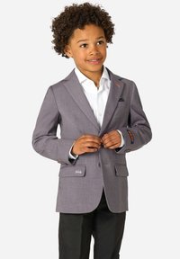 Chłopiec z kręconymi włosami zapina szary żakiet na białą koszulę, ma na sobie czarne spodnie, uśmiecha się i lekko patrzy na bok.
