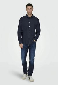 Ung mann står med en hånd i lommen iført en mørk marineblå skjorte med knapper, blå jeans og hvite sneakers foran en ensfarget bakgrunn.