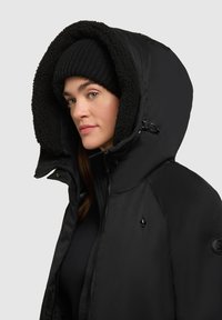 Jachetă neagră căptușită cu glugă mare căptușită cu blană și fermoar. Are un logo subtil pe piept și o șapcă de tricot neagră cu bordură.