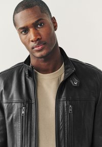 Jeune homme aux cheveux courts portant une chemise beige et une veste en cuir noire, regardant directement la caméra avec une expression neutre.