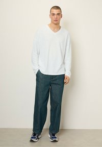 Balta ilgos rankovės V formos kaklo marškiniai su laisvu siluetu, derinami su laisvais tamsiai mėlynos spalvos džinsais ir sportiniais batais. Neutrali fono spalva.