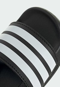 Musta liukas sandaali, jossa on kolme leveää valkoista vaakaraitaa hihnassa ja teksturoitu pohja.