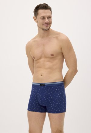 Hombre sonriente sin camiseta, con calzoncillos bóxer azules con estampado, de pie con las manos detrás de la espalda, mirando hacia su izquierda sobre un fondo liso.