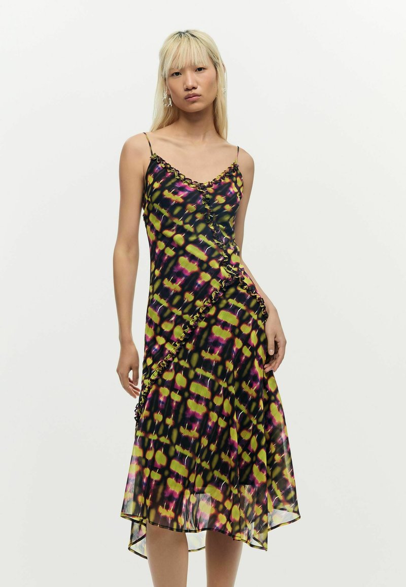 Femeie cu păr blond drept, purtând o rochie midi fără mâneci, cu model negru, galben și mov, cu detaliu de volane, pe fundal simplu.