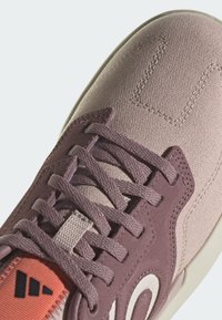 Scarpa con una tomaia in tela viola chiaro, superficie testurizzata, lacci piatti e un accento arancione-rossastro in contrasto. Presenta un dettaglio del logo bianco.