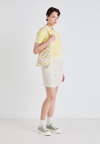 Žuta rebrasta polo majica, bež lanene šortse i zeleni tenisice s bijelim đonom. Model nosi torbu od kukičanja s cvjetnim uzorkom.