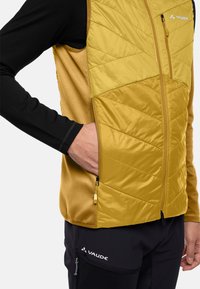 Gelbe isolierte Weste mit gestepptem Design und einem Reißverschlussfach. Darunter ein schwarzes Langarmshirt. Langlebiges Material mit glatter Textur.