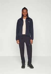 Navy fleece jacket with zip, long sleeves, and front pockets paired with black pants featuring zippered pockets. Black hiking boots complete the look.