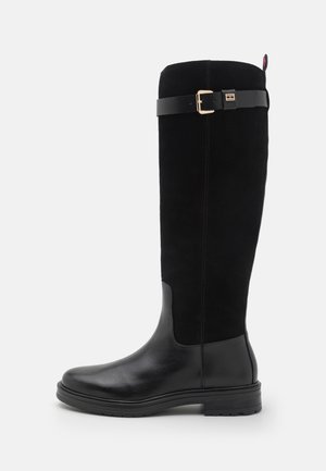 Bottes noires montantes jusqu'au genou avec pied en cuir lisse, tige en daim, accent de boucle dorée sur une sangle et semelle en caoutchouc pour l'adhérence.