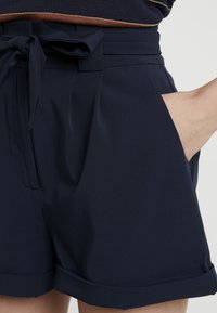 Tmavě modré kraťasy s vysokým pasem a záhyby vpředu, s ohrnutými lemy, bočními kapsami a zavazovací stuhou, nošeny s pruhovaným tričkem.