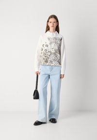 Felpa bianca con grafiche di leopardo, abbinata a jeans a gamba larga di un azzurro chiaro. Gli accessori includono una borsa nera. Fit semplice e rilassato.