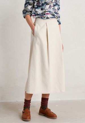 Personne portant une jupe midi crème avec un pli devant, une chemise manches longues à motif, des chaussettes bordeaux et des chaussures en cuir marron, debout sur un sol gris.