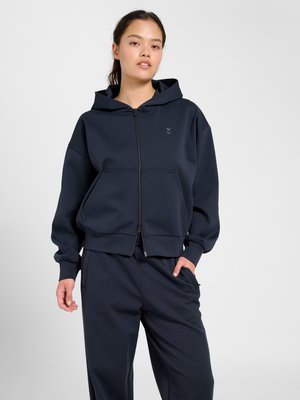 Mannequin femme portant un sweat à capuche noir zippé avec capuche et pantalon noir assorti, debout avec une main dans la poche sur fond blanc.