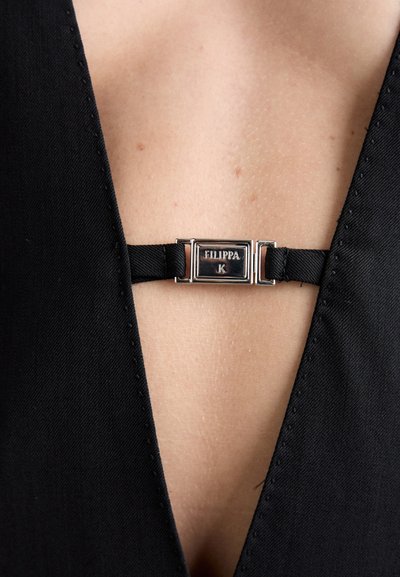 Schwarzes Kleidungsstück mit einem tiefen V-Ausschnitt. Ein Satinriemen mit einem silbernen Hardwareverschluss zeigt den Markennamen "Filippa K."