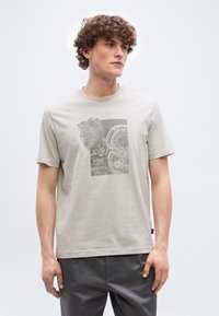 Νεαρός άνδρας με σγουρά μαλλιά που φοράει ανοιχτό μπεζ μπλουζάκι με σκούρο γραφικό δακτυλίου δέντρου και σκούρο παντελόνι, στέκεται μπροστά σε απλό φόντο.