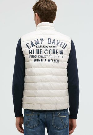Hombre con chaleco acolchado blanco con mangas largas azules y texto azul "Camp David Blue Crew" en la espalda, combinado con vaqueros azules.