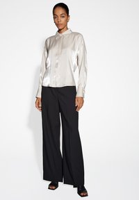 Camicia button-up in satin grigio chiaro con maniche lunghe, abbinata a pantaloni larghi neri e sandali neri con punta aperta. Linee pulite e texture liscia.