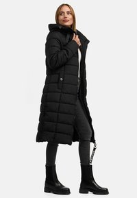 Manteau long noir rembourré avec une capuche, des détails de fermeture éclair et de boutons. Porté sur un jean gris et des bottes noires, avec un extérieur texturé.
