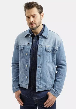 Mężczyzna z krótkimi włosami i brodą, ubrany w jasnoniebieską kurtkę dżinsową na ciemnoniebieską koszulę, stojący na jednolitym tle.