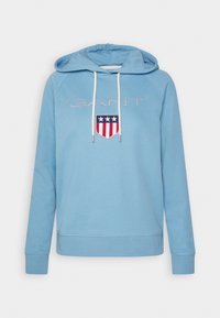 Sweat à capuche bleu clair avec cordons blancs, texte brodé "GANT" et logo écusson rouge, blanc et bleu avec étoiles et rayures sur la poitrine.