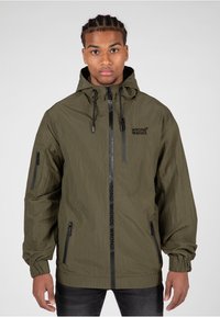 Olivegrüne leichte Jacke mit Kapuze, Reißverschluss vorne und Seitentaschen. Mit Logo-Druck entlang des Reißverschlusses vorne und elastischen Bündchen.