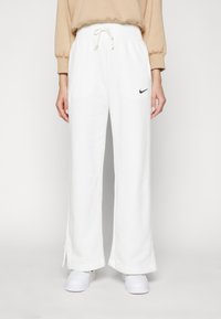 Hvite joggebukser med snøring i livet, bred benvegg, sideskår i kanten og en liten svart Nike-logo på venstre foran.