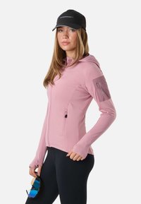 Frau in einem rosa Kapuzenpullover mit Reißverschluss und schwarzer Mütze, die blaue Sportsonnenbrillen mit Tönung hält und vor einem schlichten weißen Hintergrund steht.
