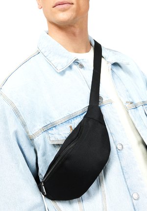 Bolsa de malla negra tipo bandolera con un compartimento con cremallera, llevada cruzada sobre el cuerpo, combinada con una chaqueta de mezclilla azul claro. Correa ajustable para mayor comodidad.