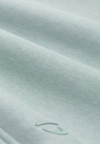 Lichtblauwe katoenen stof met een gladde textuur, met subtiele plooien en een klein, reliëf logo in de rechterbenedenhoek.