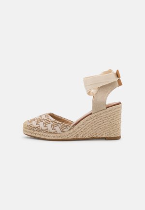 Beigen värinen kiilakorkoinen espadrillos-kengän, jossa on kudottu kuvio, nilkan ympärille kääritettävä hihna ja punottu juuttikorko, yksinkertaisella valkoisella taustalla.