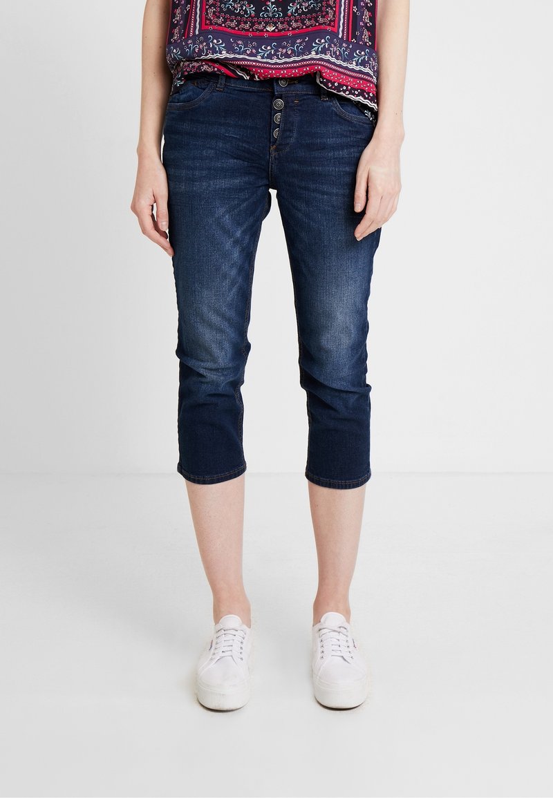 Tmavě modré capri džíny z denimu, s pěti knoflíky na zapínání, předními kapsami a ohrnutými nohavicemi. Spárované s bílými sportovními botami.