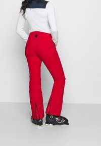 Rode skibroek met ritsen bij de enkels, gecombineerd met zwarte skischoenen. Het materiaal lijkt stevig en is ontworpen voor wintersport.